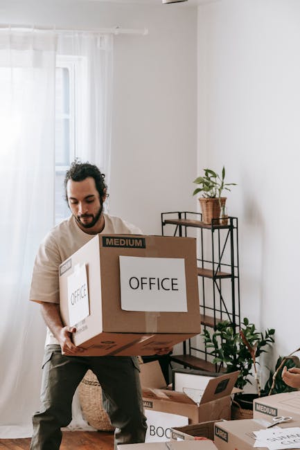 A smiling man and woman are standing indoors, holding a large cardboard moving box together during a home relocation process. Behind them, there are several stacked cardboard boxes of varying sizes, s