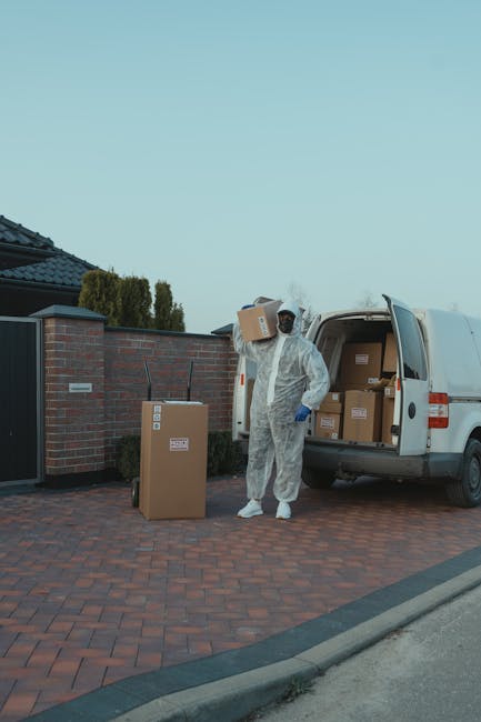 Two men engaged in a home relocation process on Brockley Road, loading cardboard boxes into a white cargo van parked on a paved driveway. One man is pushing a hand truck carrying a large cardboard box