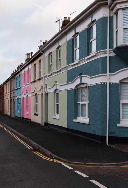 A row of colorful terraced houses in Brockley featuring pastel shades of blue, pink, green, and beige, with white window frames and decorative mouldings. The photograph is taken from the pavement, sho