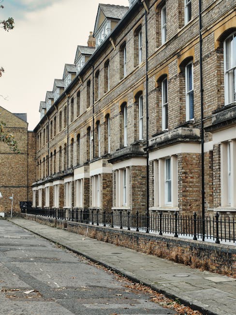 A row of colorful terraced houses in Brockley featuring pastel shades of blue, pink, green, and beige, with white window frames and decorative mouldings. The photograph is taken from the pavement, sho