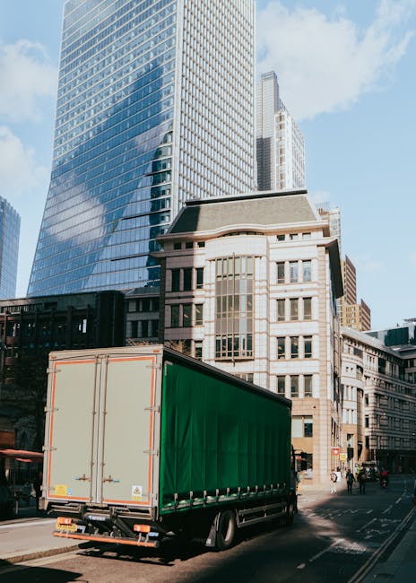 A rear view of a large, silver box truck with its back doors open, parked in an alleyway beside a brick building. The truck's interior is visible, showing scattered packing materials and miscellaneous