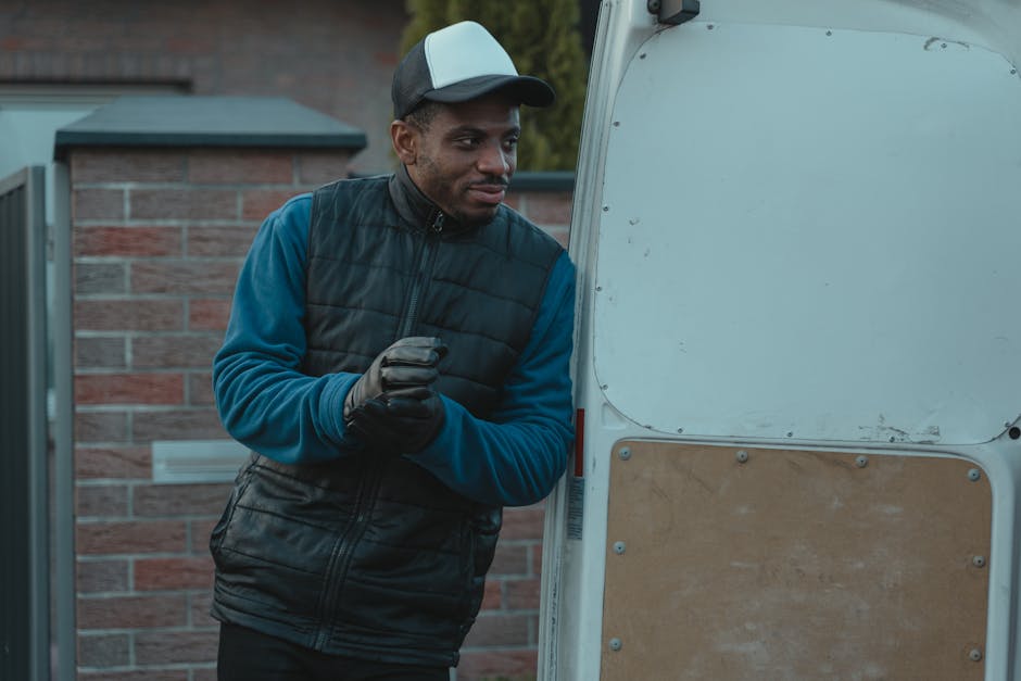 A smiling man with curly black hair and a beard is sitting inside the empty cargo area of a white van, which is parked outdoors with its rear doors wide open. He is wearing a dark blue t-shirt with a