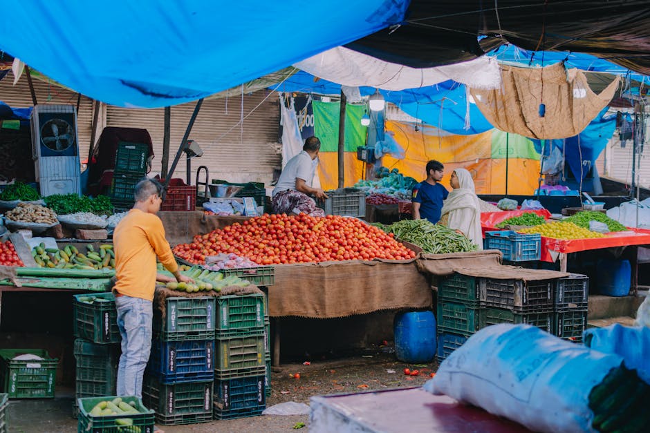 A busy outdoor market stall under a makeshift tent made from blue, white, and beige tarpaulins, featuring plastic crates and wooden tables loaded with fresh produce such as tomatoes, cucumbers, and green leafy vegetables. A man in an orange top is stacking or arranging vegetables, while a woman in a white shirt faces away from the camera, possibly managing the stall. A young girl in a blue t-shirt stands nearby, observing the scene. In the background, a person dressed in a white top and patterned skirt is attending to the produce. The ground is dirt, and various plastic bags and packaging materials are visible. The scene is well-lit, suggesting daytime, with natural light filtering through the tent coverings. This image captures the typical setting of a marketplace, relating to home relocation or packing and moving services provided by Man and Van Brockley, who may assist with storage or transport of household goods after purchase or sale.
