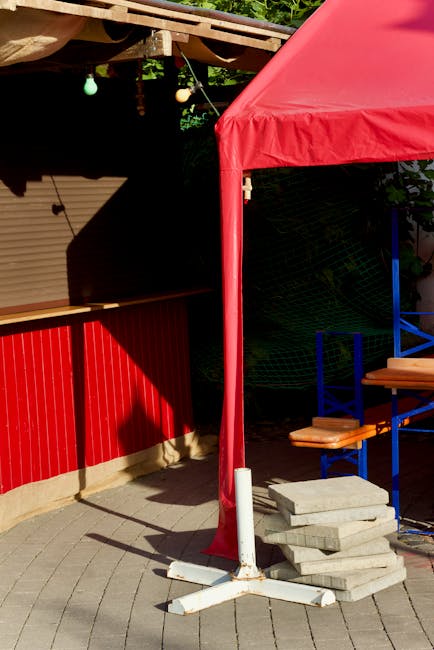 A red canopy tent with fabric sides and metal poles, set up on a paved outdoor area adjacent to a red wooden structure, appears to be part of an organized event or market stall. There are several stacked paving stones or concrete slabs placed on the ground near the tent's support pole, which is stabilized with a white base. Behind the tent, there are blue chairs with wooden seats, likely part of seating arrangements. The background features green foliage and string lights hanging from a wooden overhead structure, suggesting a semi-outdoor environment. The scene depicts a stable area for ongoing activities related to house removals, packing, or setting up a market stall, which aligns with services provided by Man and Van Brockley in the context of home relocation and furniture transport.