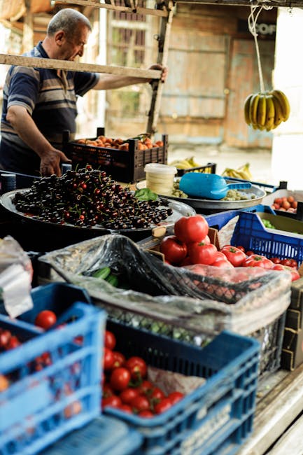 A busy outdoor market stall under a makeshift tent made from blue, white, and beige tarpaulins, featuring plastic crates and wooden tables loaded with fresh produce such as tomatoes, cucumbers, and green leafy vegetables. A man in an orange top is stacking or arranging vegetables, while a woman in a white shirt faces away from the camera, possibly managing the stall. A young girl in a blue t-shirt stands nearby, observing the scene. In the background, a person dressed in a white top and patterned skirt is attending to the produce. The ground is dirt, and various plastic bags and packaging materials are visible. The scene is well-lit, suggesting daytime, with natural light filtering through the tent coverings. This image captures the typical setting of a marketplace, relating to home relocation or packing and moving services provided by Man and Van Brockley, who may assist with storage or transport of household goods after purchase or sale.