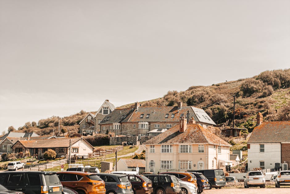 A view of a residential street showing a parking area filled with various compact cars and vans in front of a row of houses with different architectural styles. Closer to the foreground, there are a few vehicles including a black van and a white car parked on the pavement. Behind the parked vehicles, several houses with pitched roofs and chimneys are visible, including a cream-colored semi-detached house with white-framed windows and a brown tiled roof. Further in the background, more residential buildings sit on a hillside, including larger modern-style homes with large windows, some with glass balconies, and terraced houses. The hillside is covered with bushes and grass, and the sky above is mostly clear with a few clouds. This scene exemplifies the typical environment for home relocation and furniture transport services offered by Man and Van Brockley, highlighting the outdoor parking and housing context involved in house removals in the Hilly Fields, Brockley area.